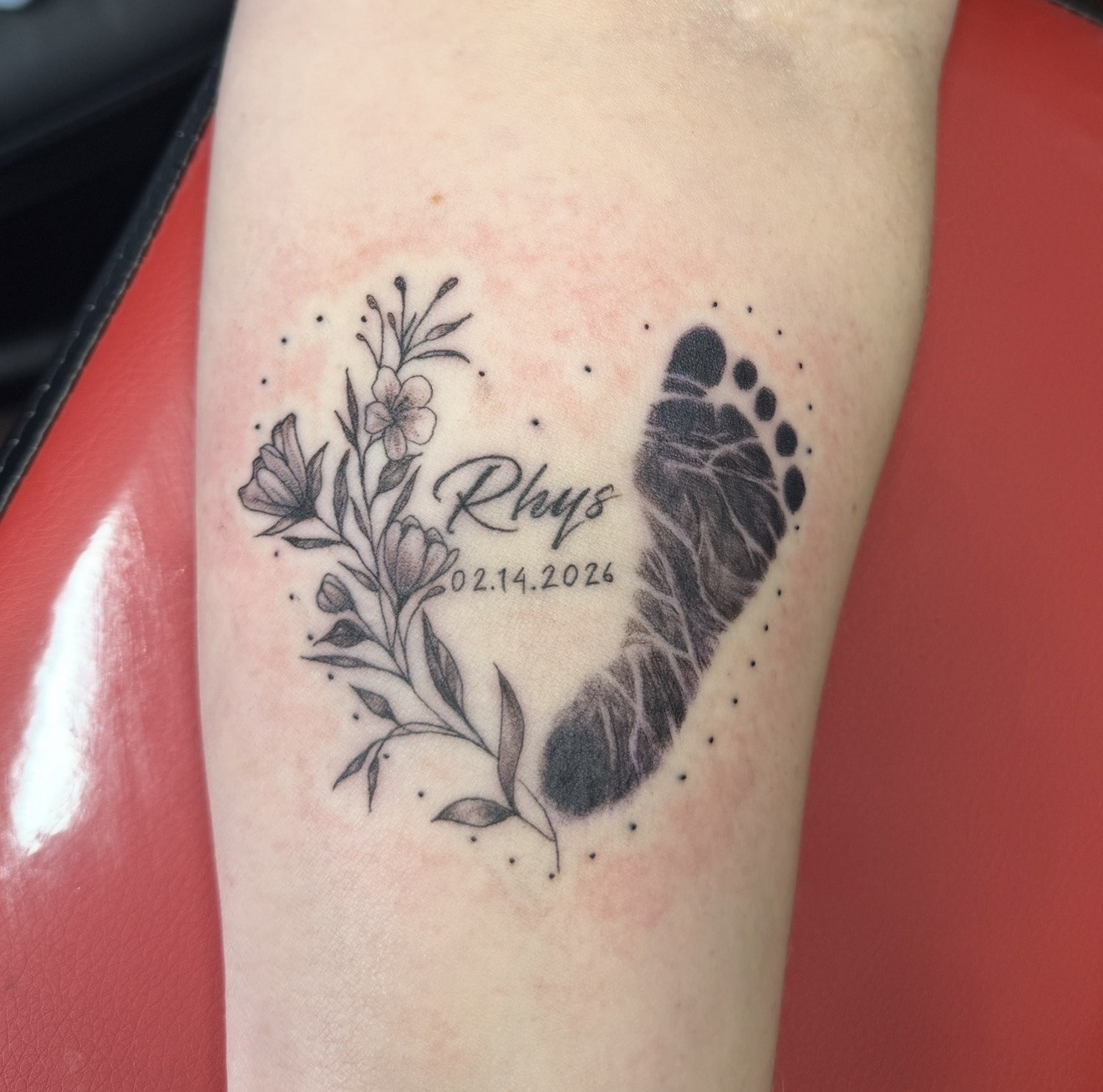 A footprint framed by delicate flowers, with a name and date in lettering. Tattoo work by Joseph Rojas.
